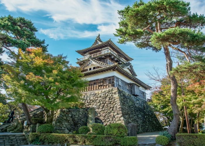 Looking up at Maruoka Castle, surrounded by trees and blue sky. The leaves are started to take on the colors of fall.